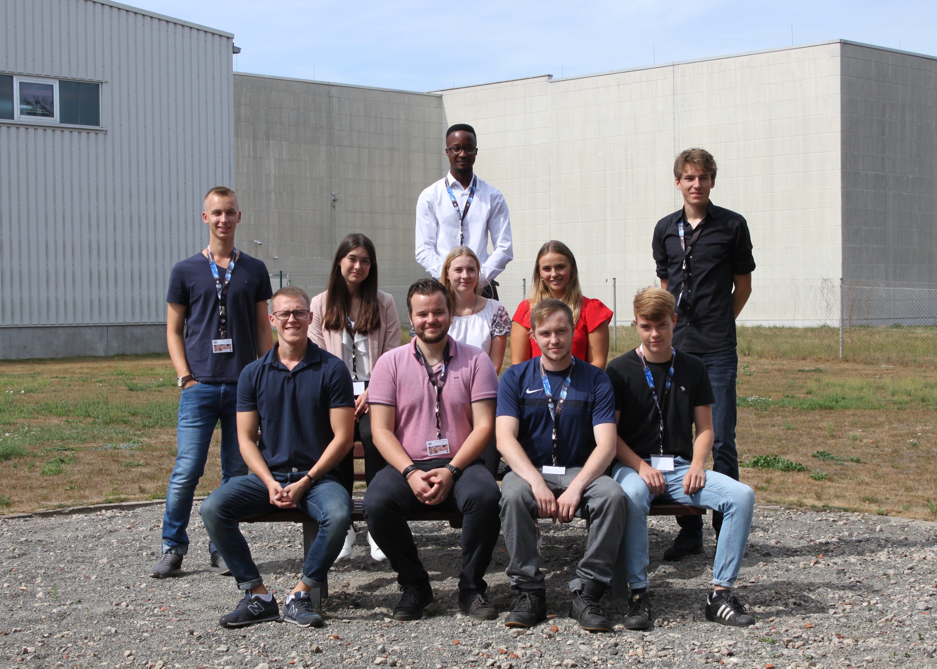 Eine Gruppe von Personen mit Lanyards steht vor einem großen grauen Gebäude und einige sitzen auf einer Bank.