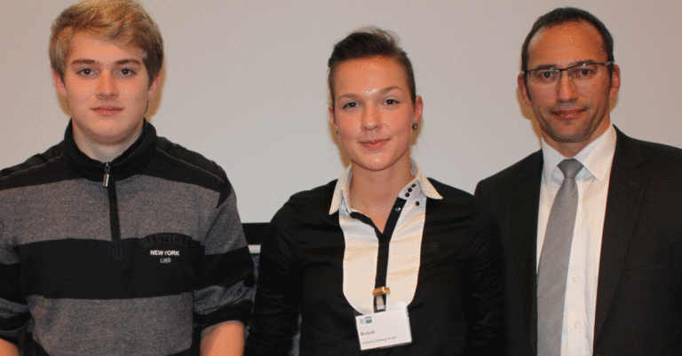 Ein blonder junger Mann, eine brünette Frau und ein Mann in Anzug und mit Brille stehen nebeneinander und blicken in die Kamera.