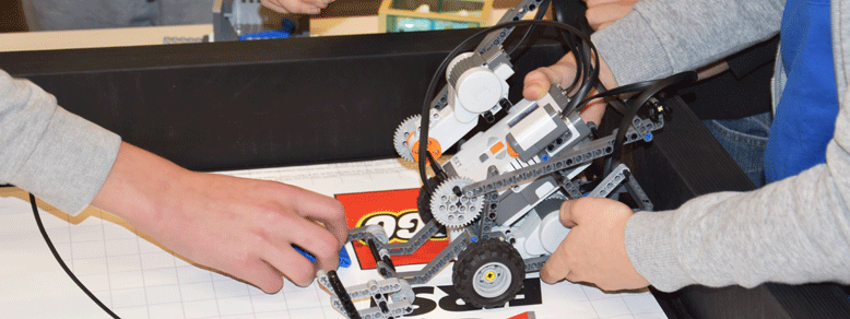 Rechts stellt eine Person stellt einen Roboter aus Lego auf einen Tisch mit Lego-Logo und von links greift jemand mit einer Hand nach dem vorderen Bauteil des Roboters.