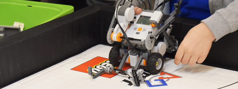 Auf einem Tisch mit Lego-Logo steht ein Roboter aus Lego, der von einer Person mit grauem Oberteil dort hingestellt wird.