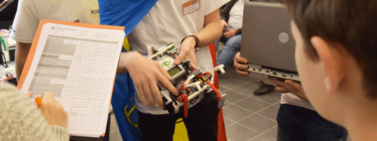 Ein Junge in weißem T-Shirt hält einen Roboter aus Lego und rechts hakt eine Person auf einem Klemmbrett etwas ab.