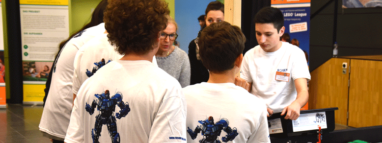 Mehrere Personen in weißen T-Shirts mit einem Rückenaufdruck eines Roboters stehen an einem Tisch und im Hintergrund redet eine blonde Frau mit Brille, während alle nach unten auf den Tisch schauen.