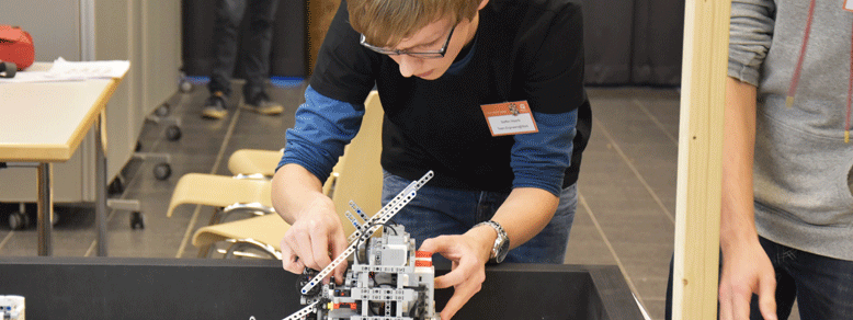 Ein Junge mit dunkler Kleidung und blauer Jeans und Brille beugt sich nach vorne und hält mit beiden Händen einen Roboter aus Lego fest.
