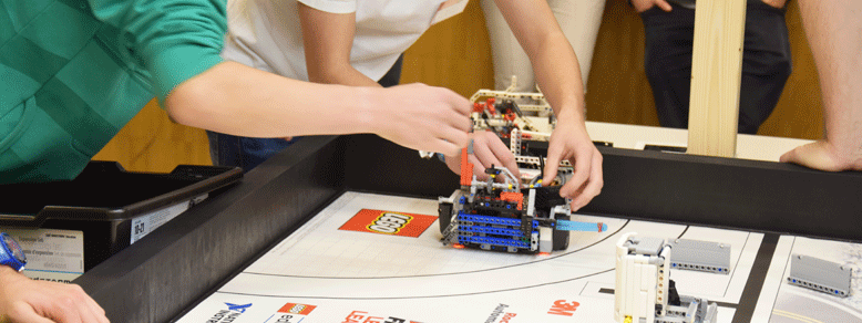 Mehrere Personen beugen sich über einen Tisch mit Legoteilen und einem Roboter aus Lego, zwei Leute auf der linken Seite berühren den Roboter mit ihren Händen.