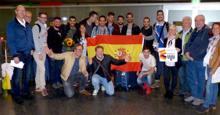 Eine Gruppe von Menschen vom Projekt von PractiGo stehen zusammen am Flughafen mit Koffern und mittig in der Gruppe wird die spanische Flagge von zwei Personen gehalten.