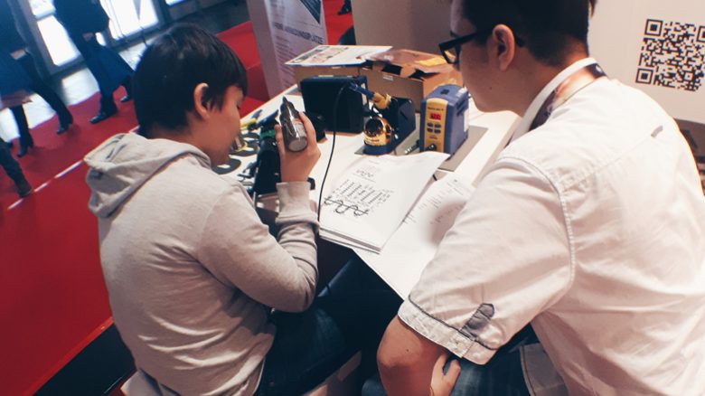 Auf der Jobmesse sitzt ein Junge an einem Tisch und lötet bei einem Lötprojekt, während ein Auszubildender neben ihm sitzt und unterstützt.