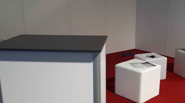 Vor einer weißen Wand und rotem Teppichboden stehen zwei weiße Hocker, auf denen Kabel und Papiere liegen und ein weißer Messecounter mit schwarzer Tischplatte.