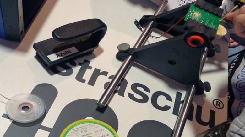 Auf einem Tisch liegen verschiedene Materialien für das Lötprojekt und in der oberen rechten Ecke lötet jemand an einer Platine.