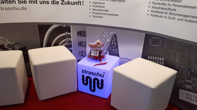 Vor einer Messewand stehen drei weiße Hocker und ein beleuchteter Kasten mit straschu Logo und einer Glasschale mit Giveaways.