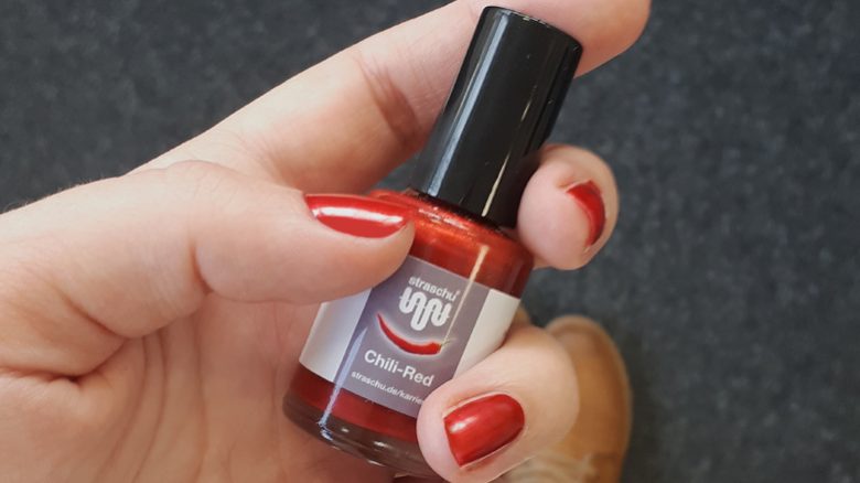 Von einer Hand mir rot lackierten Fingernägeln wird eine Nagellackflasche mit rotem Lack und grauem Etikett mit straschu Logo gehalten.
