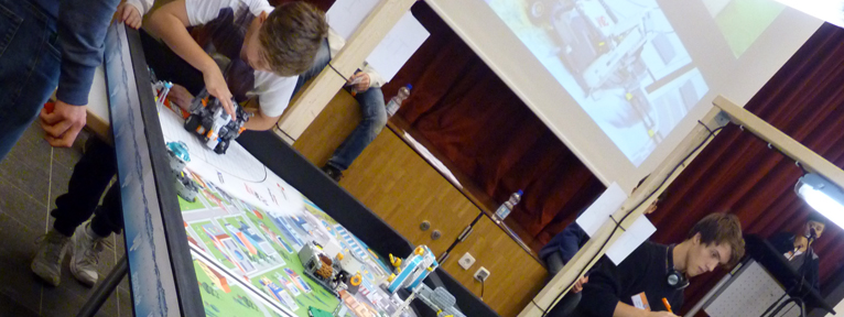Bei der First Lego League stellt ein Junge einen Legoroboter auf einen Tisch, auf dem mehrere Konstruktionen aus Lego stehen.