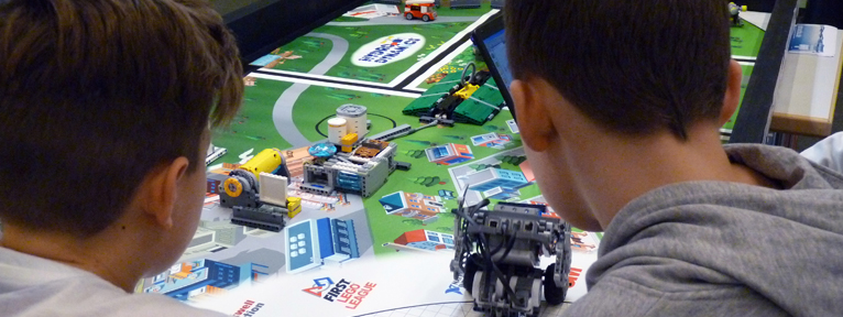 Zwei Jungs schauen auf einen Tisch der First Lego League mit einem Roboter aus Lego.