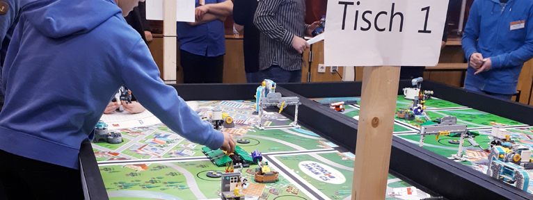 Zwei Tische mit Lego stehen nebeneinander, an dem vorderen Tisch ist ein Holzstab mit einem Papier mit Schriftzug "Tisch 1" befestigt und ein Junge greift nach einem Legokonstrukt auf dem Tisch.