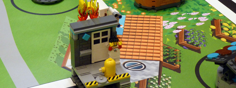 Mittig im Bild ist eine Legokonstruktion, die eine in Flammen stehende Hauswand mit Bewohner vor der Haustür zeigt und an den Bildrändern sind weitere Lego-Bauten auf einer grünen Unterfläche mit Garten-Motiven.