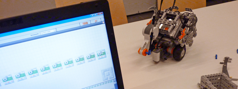 Auf der linken Bildseite ist ein Teil von einem Laptopbildschirm und auf der rechten Bildseite steht ein Lego-Roboter und Teil einer weiteren Konstruktion aus Lego auf einem hellen Tisch.