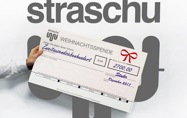 Eine Hand hält einen Check für eine Weihnachtsspende vor das straschu Logo im Hintergrund.