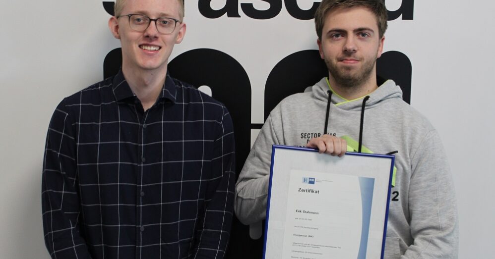 Zwei Männer, die am Projekt Energyscouts mitgewirkt haben, stehen vor einer Wand mit straschu Logo und der rechte hält ein gerahmtes Zertifikat.