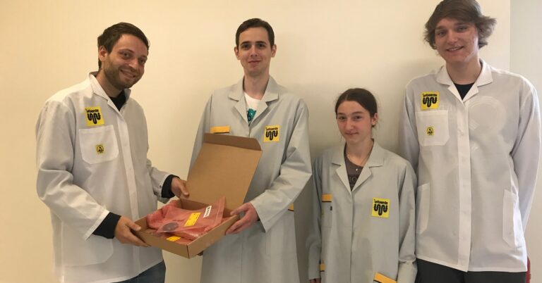 Vier Personen in hellen Kitteln mit straschu Logo als CanSat Team Starbugs und die Männer auf der linken Seite halten einen aufgeklappten Karton mit in roter Folie eingepackten Produkten