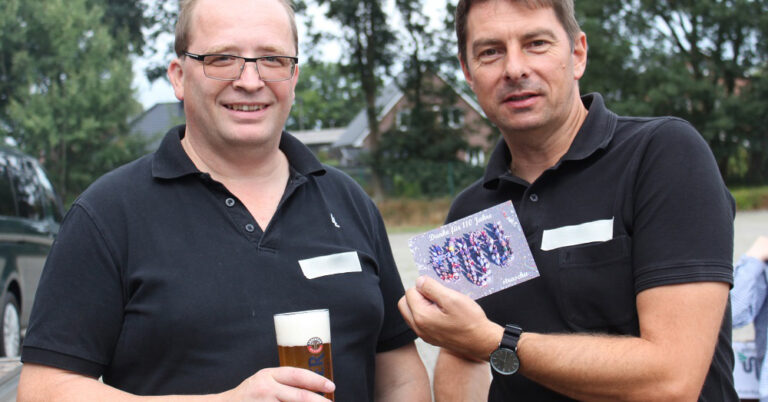Zwei Männer in dunklen Poloshirts, der linke hält in seiner Hand ein Bierglas und der rechte zeigt eine straschu -Postkarte in die Kamera