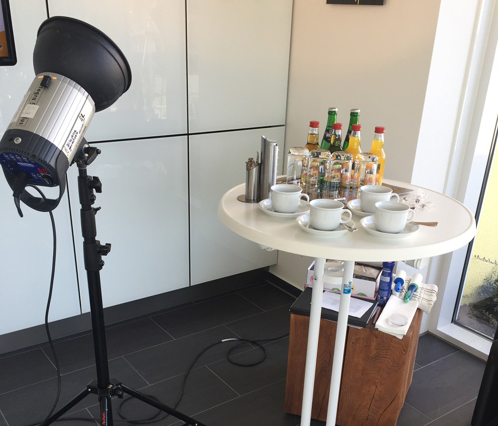 Auf einem weißen Stehtisch stehen Kaffetassen und Getränkeflaschen, im hintergrund steht ein Hocker auf dem Boden und links steht eine Lampe.