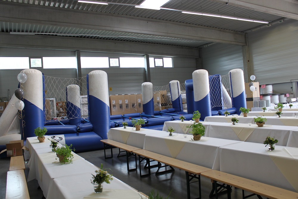 In einer großen Halle stehen im Vordergrund lange Tischreihen, die mit weißen Tischdecken eingedeckt sind und im Hintergrund ist eine weißblaue Trampolinlandschaft aufgebaut.