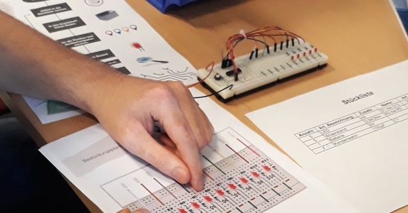 Auf einem Tisch liegen Unterlagen und ein Schaltgerät für den Berufsparcours der BBs Syke und eine Person zeigt mit einem Finger auf eine der Unterlagen.