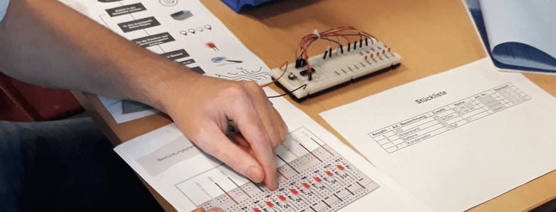 Auf einem Tisch liegen Unterlagen und ein Schaltgerät für den Berufsparcours der BBs Syke und eine Person zeigt mit einem Finger auf eine der Unterlagen.