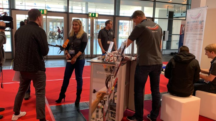 Der straschu Stand auf der Jobmesse in Bremen, wo ein Mitarbeiter am Messcounter steht, zwei Jungs an einem Tisch zum Löten sitzen und rechts eine straschu Mitarbeiterin von einem ARD-Fernsehteam fürs TV interviewt wird.