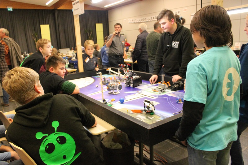 Bei der First Lego League steht der straschu Tisch mit Legos zur Raumfahrt und Schüler stehen und sitzen um den Tisch herum.
