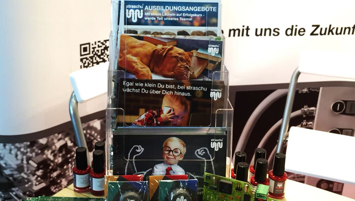 Ein Kartenaufsteller, mehrere rote Nagellackflaschen und eine grüne Leiterplatte beim Messestand auf der vocatium.