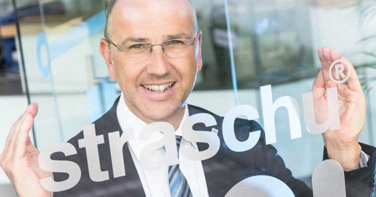 Ein Mann mit Brille steht vor einer Glasscheibe, auf der straschu steht, und hält seine Hände an den Schriftzug und lächelt.