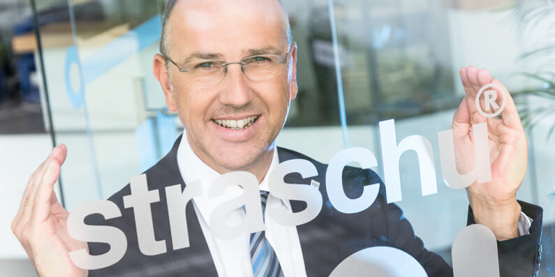 Ein Mann mit Brille steht vor einer Glasscheibe, auf der straschu steht, und hält seine Hände an den Schriftzug und lächelt.