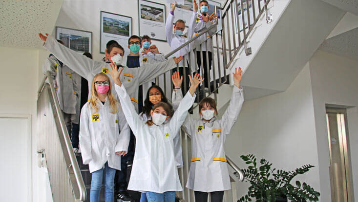Eine Gruppe von Kindern in weißen Kitteln steht auf einer Treppe und streckt die Arme nach oben.