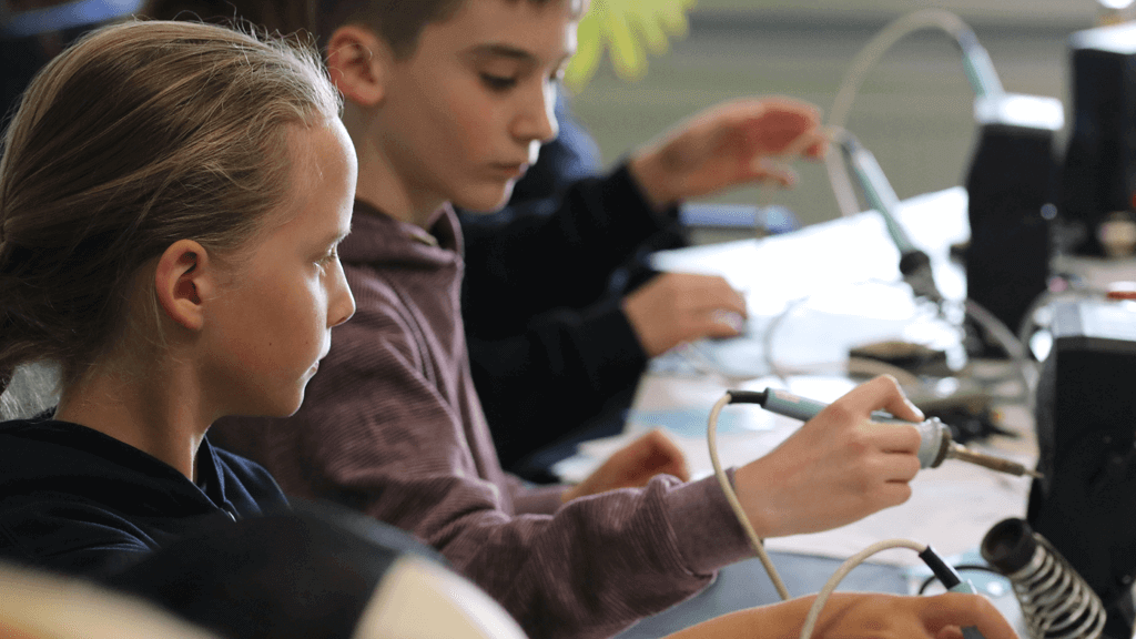Eine Gruppe von Kinder löten mit Lötkolben in der Hand am Zukunftstag ihr eigenes Lötprojekt.