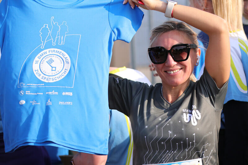 Eine Frau in einem straschu-Shirt und mit Sonnenbrille hält ein blaues T-Shirt mit einem Schriftzug vom Firmenlauf in die Kamera und lächelt.