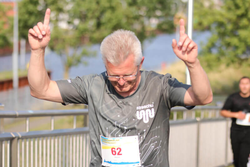 Ein Mann in straschu-Shirt läuft beim Firmenlauf mit und streckt beide Hände nach oben.
