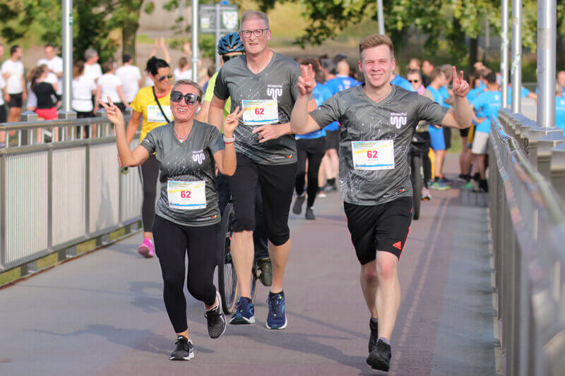Mehrere Menschen mit straschu-Shirts laufen beim Firmenlauf über eine Brücke und lächeln.