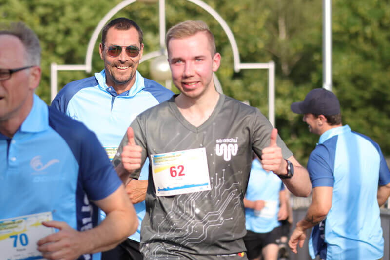 Ein Mann mit straschu-Shirt läuft beim Firmenlauf mit und zeigt beide Daumen nach oben.
