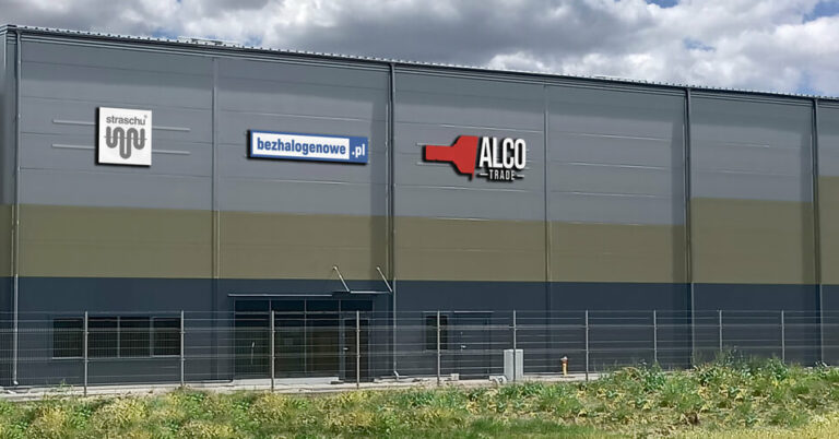 Ein dunkles Gebäude mit straschu Logo und einem Zaun und Büschen davor.