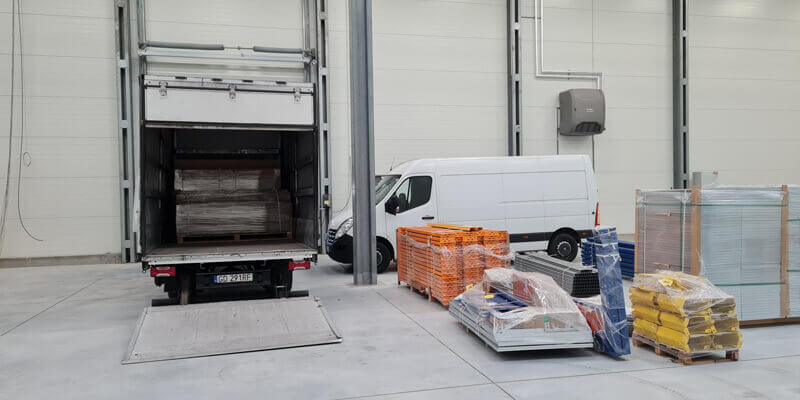 Ein offener Lieferwagen und ein Sprinter stehen in einer Halle und im Bildvordergrund sind Artikel auf Paletten eingepackt und eingewickelt in Packfolie.
