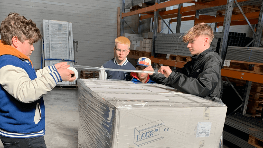 Zwei Jungs in einem Lager spannen Packfolie über ein Paket am Zukunftstag während zwei Jungs hinter den Paketen stehen und zuschauen.