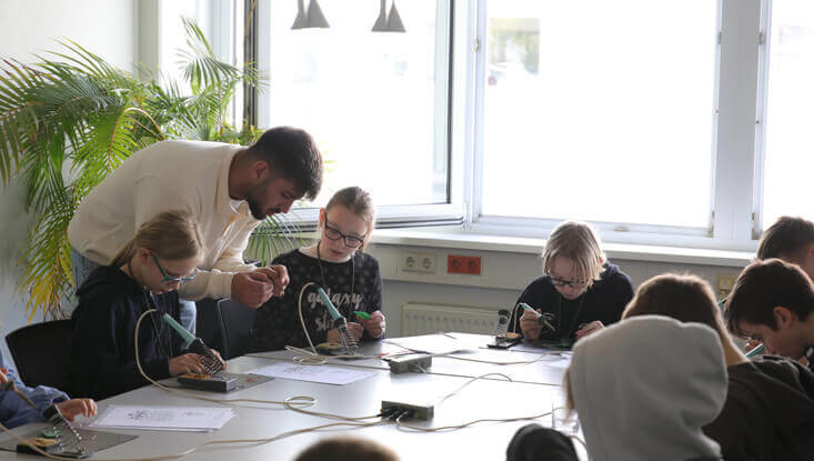 Kinder sitzen an einem Tisch und löten am Zukunftstag 2024 ihr eigenes Lötprojekt, ein Mann steht hinter dem Tisch und hilft ihnen dabei.