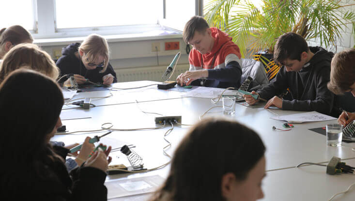 Kinder sitzen an einem Tisch und löten am Zukunftstag 2024 ihr eigenes Lötprojekt.