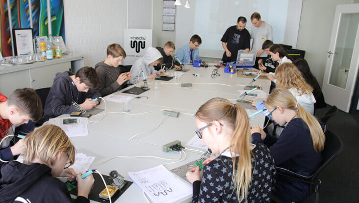Kinder sitzen an einem Tisch und löten am Zukunftstag 2024 ihr eigenes Lötprojekt, vorne vor den Tischen stehen zwei Männer und helfen.