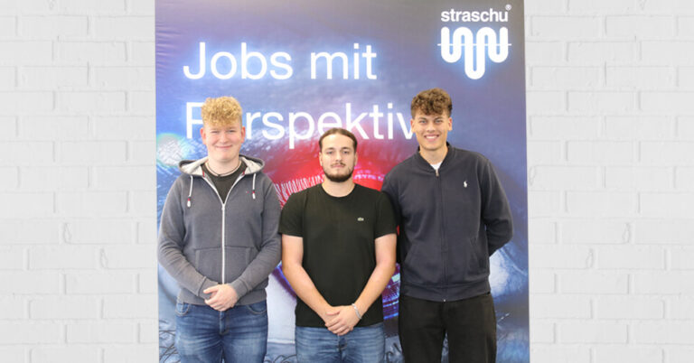drei Männer lächeln und stehen vor einem straschu-Plakat