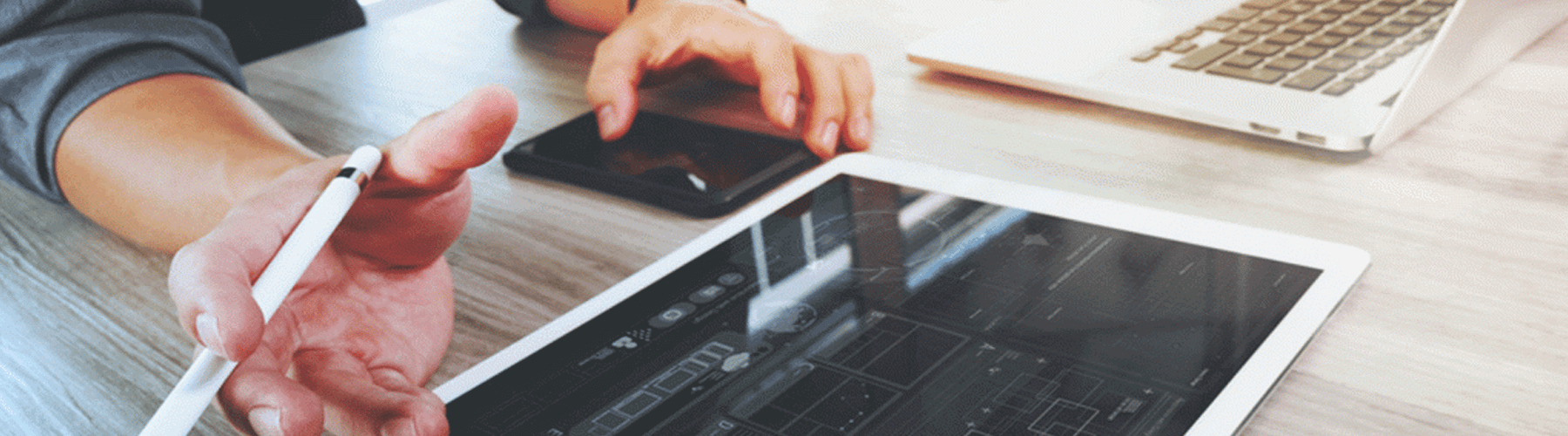 An einem Tisch sitzt eine Person und swipt auf einem Handy und hält mit der anderen Hand einen Stift. Ein Tablet liegt auf dem Tisch, im Hintergrund steht ein aufgeklappter Laptop.