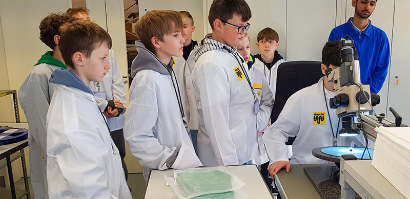 Ein Schüler im weißen Kittel schaut durch ein Mikroskop und eine Schülergruppe schaut zu am Zukunftstag.
