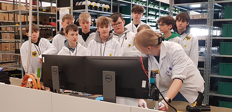 Eine Gruppe Kinder steht vor einem Arbeitsplatz und schaut zu einer Mitarbeiterin am Zukunftstag.