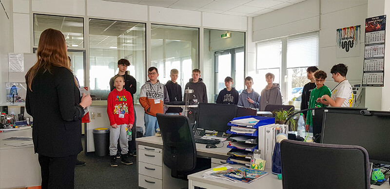 Eine Gruppe von Schülern steht in einem Büro und schaut zu einer Mitarbeiterin, die ein Prospekt erklärt am Zukunftstag.