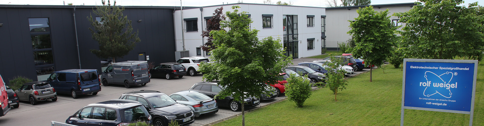 Ein Gebäude mit parkenden Autos davor, Begrünung und ein Firmenschild von rolf Weigel.